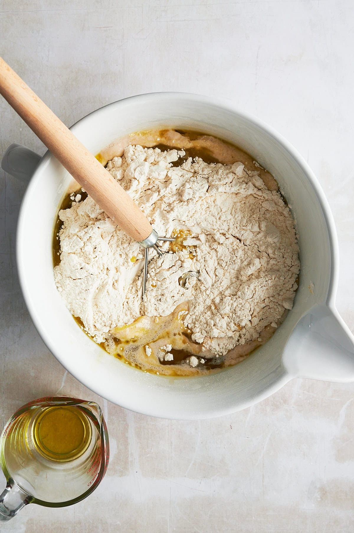 White bowl and dough beater mixing flour, olive oil for focaccia dough with glass measuring cup of olive oil.