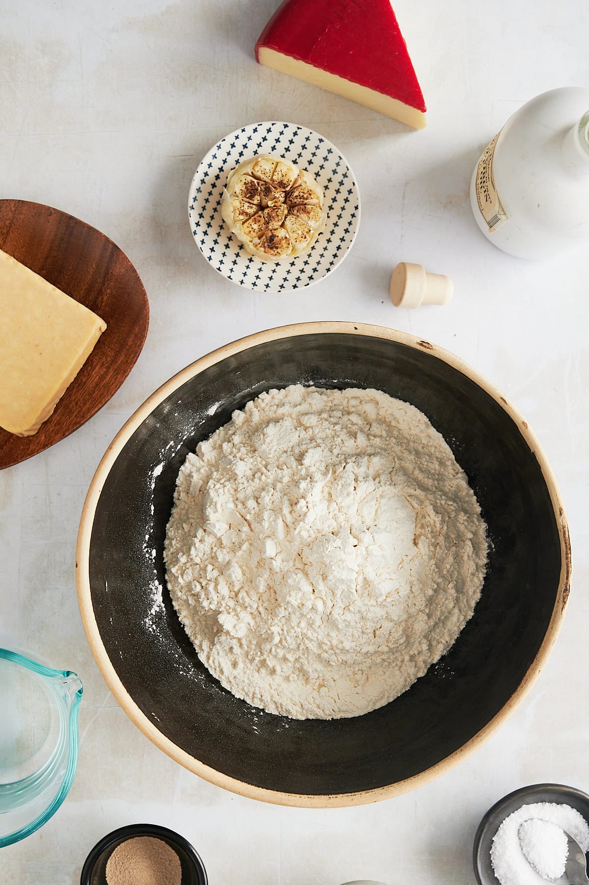 Garlic bread focaccia ingredients in bowls and plates including flour, roasted garlic, fontina cheese, yeast, olive oil, and salt.