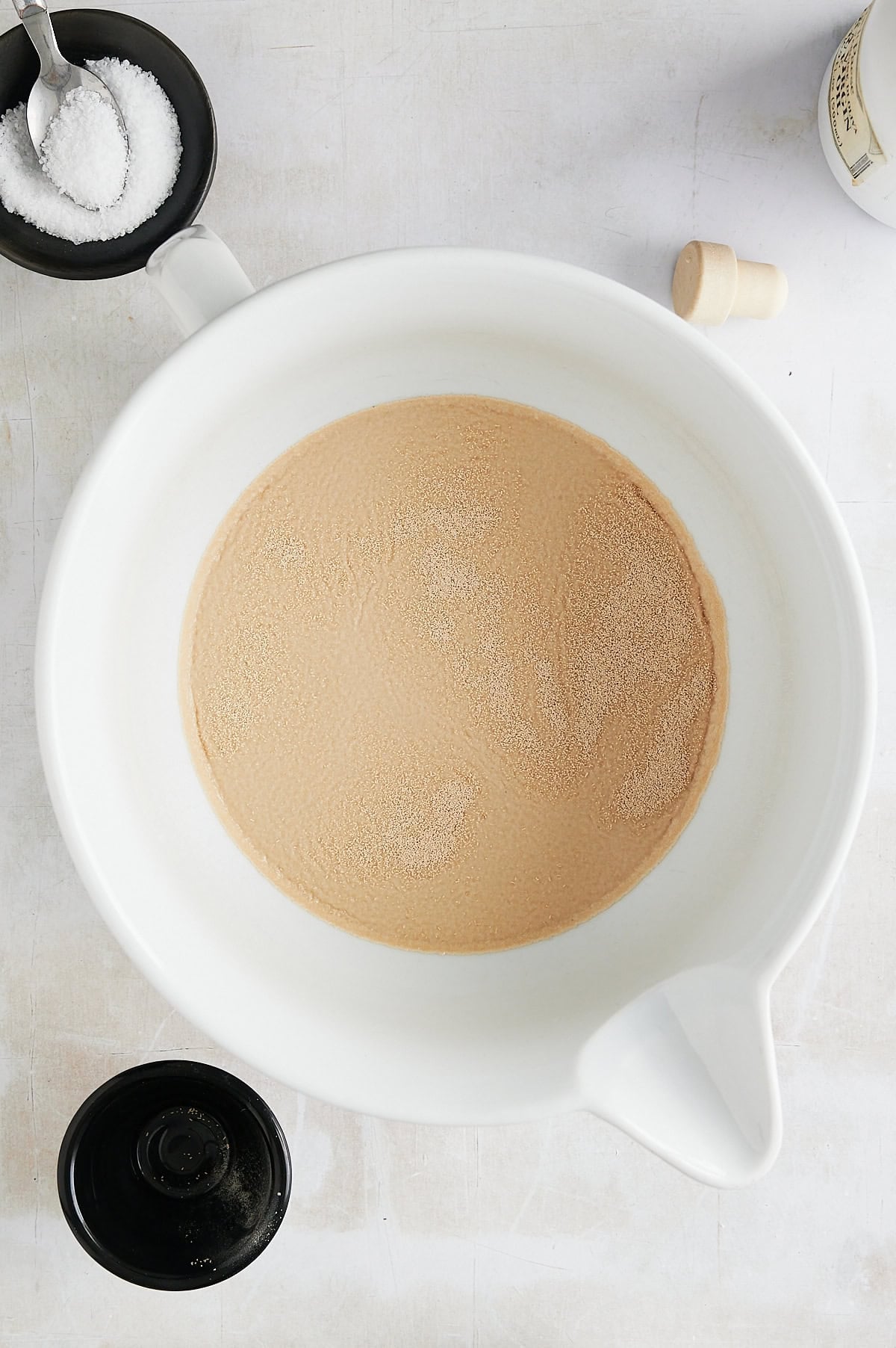 White bowl of yeast topped water with small black bowl of salt with a spoon and white bottle of olive oil.