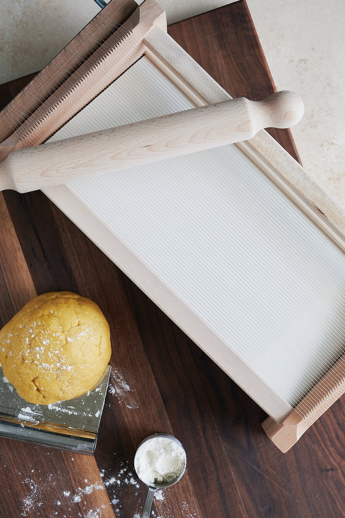 Chitarra pasta machine with a rolling pin sitting on wooden board alongside a ball of pasta dough and cup of flour.
