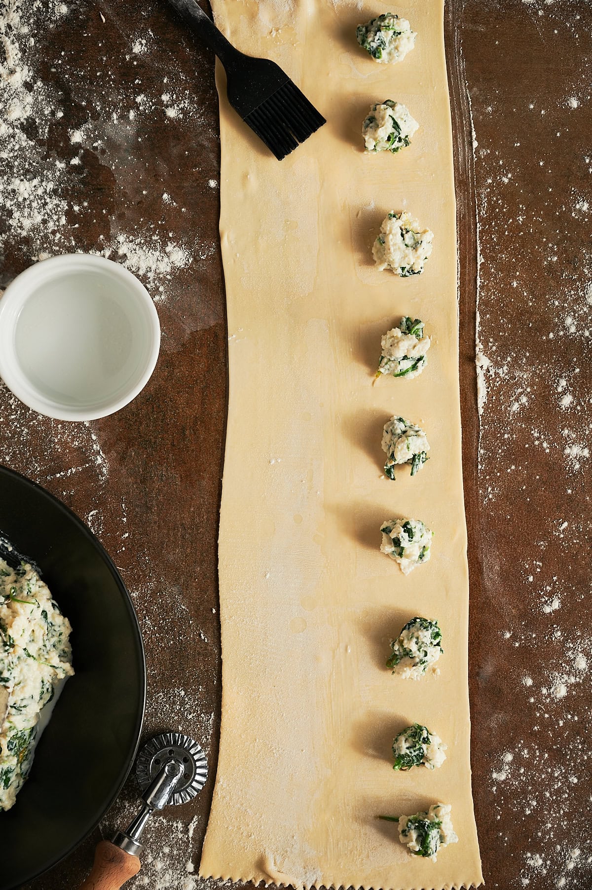 Wooden board of rolled out pasta dough with spoonfuls of spinach ravioli filling and a black brush and bowl of water.