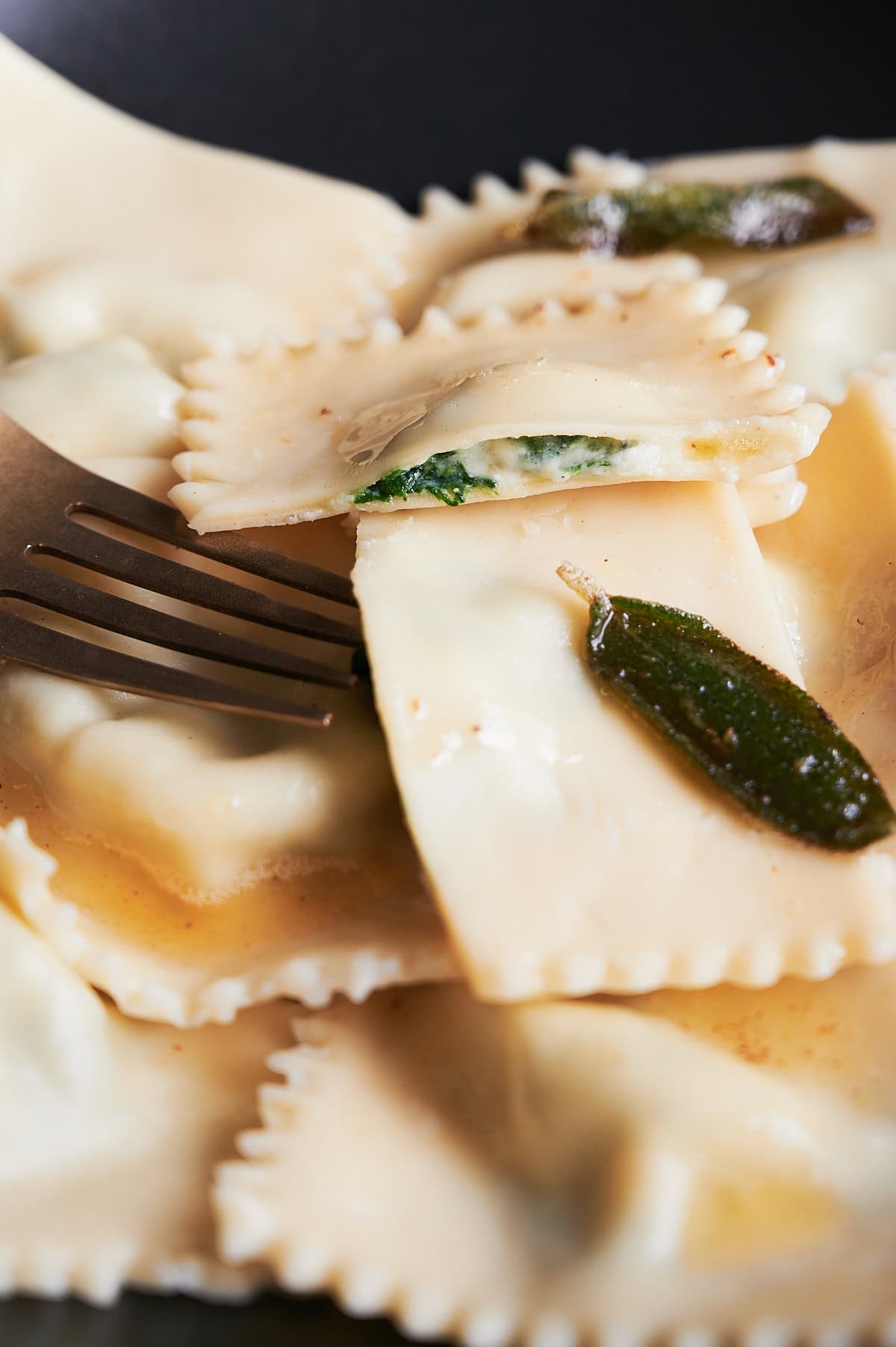 Zoom of cooked spinach ravioli with sage leaves and butter sauce and a fork.
