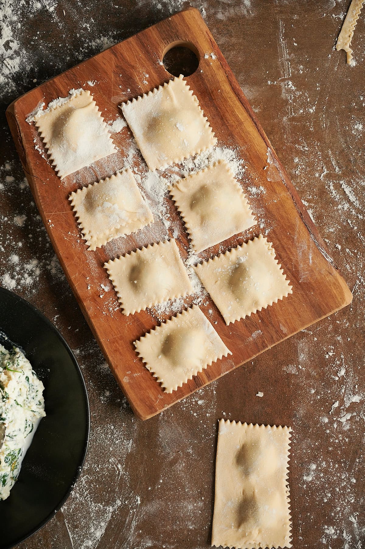 Floured wooden board with uncooked cut and rolled spinach ravioli and black bowl of fillling.