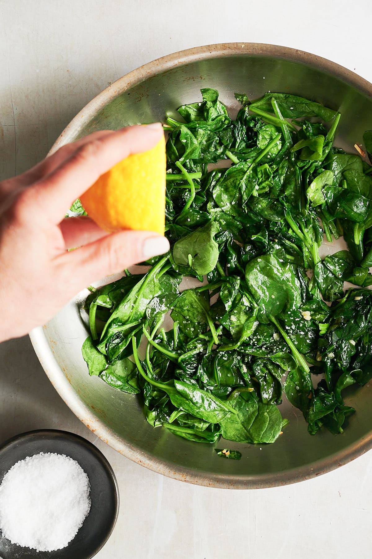 Hand squeezing lemon over stainles skillet of sauteed spinach and black bowl of kosher salt.
