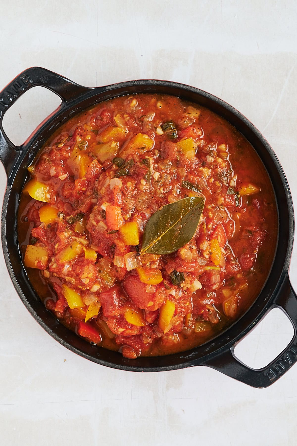 Angled black skillet of stewed tomatoes with a bay leaf on top on a cream background.