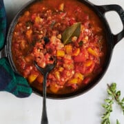 Black skillet of stewed tomatoes with a black spoon holding some and a bay leaf with a blackwatch plaid napkin and fresh oregano.