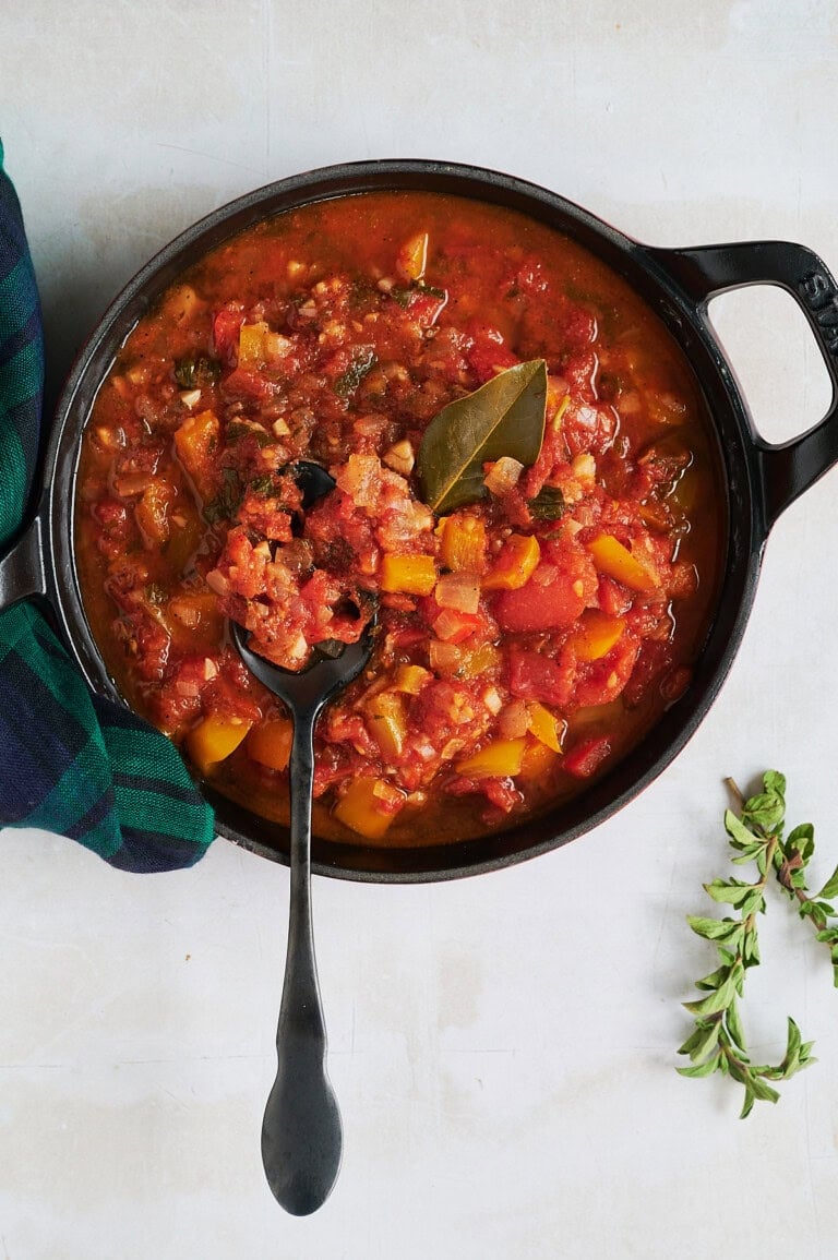Black skillet of stewed tomatoes with a black spoon holding some and a bay leaf with a blackwatch plaid napkin and fresh oregano.