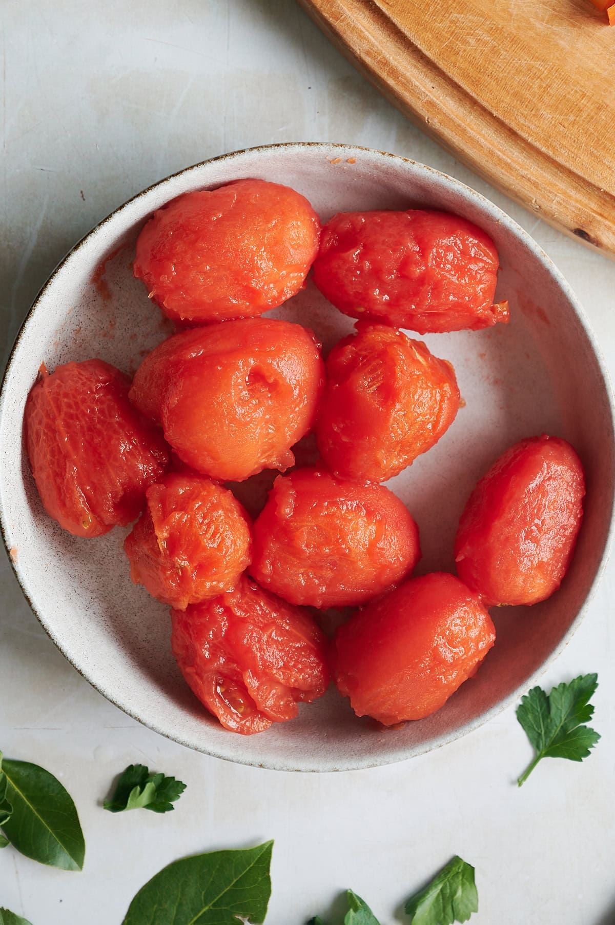 White bowl of peeled tomatoes with fresh herbs sprinkled on a cream background.