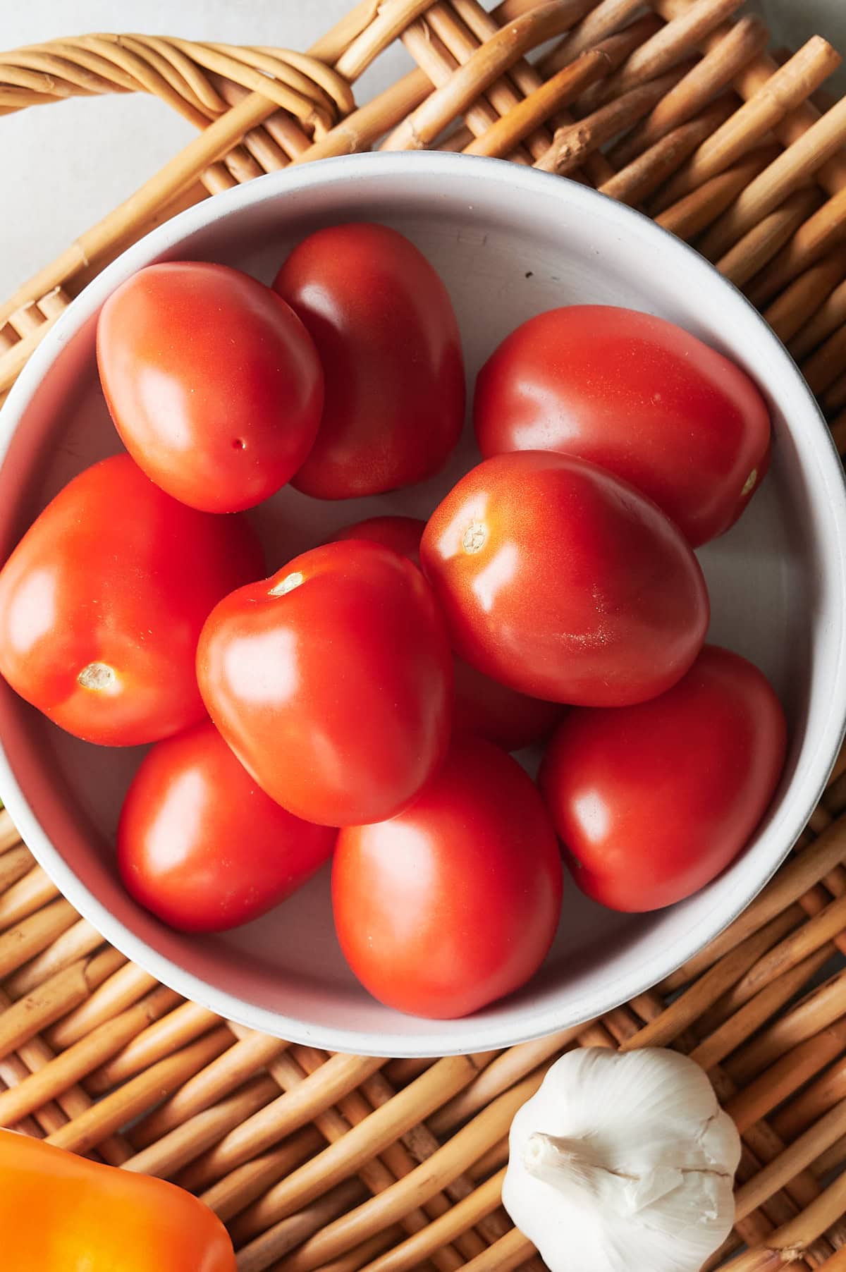 Closeup of bowl of Roma tomatoes in a white bowl on a rattan placemat with head of garlic.
