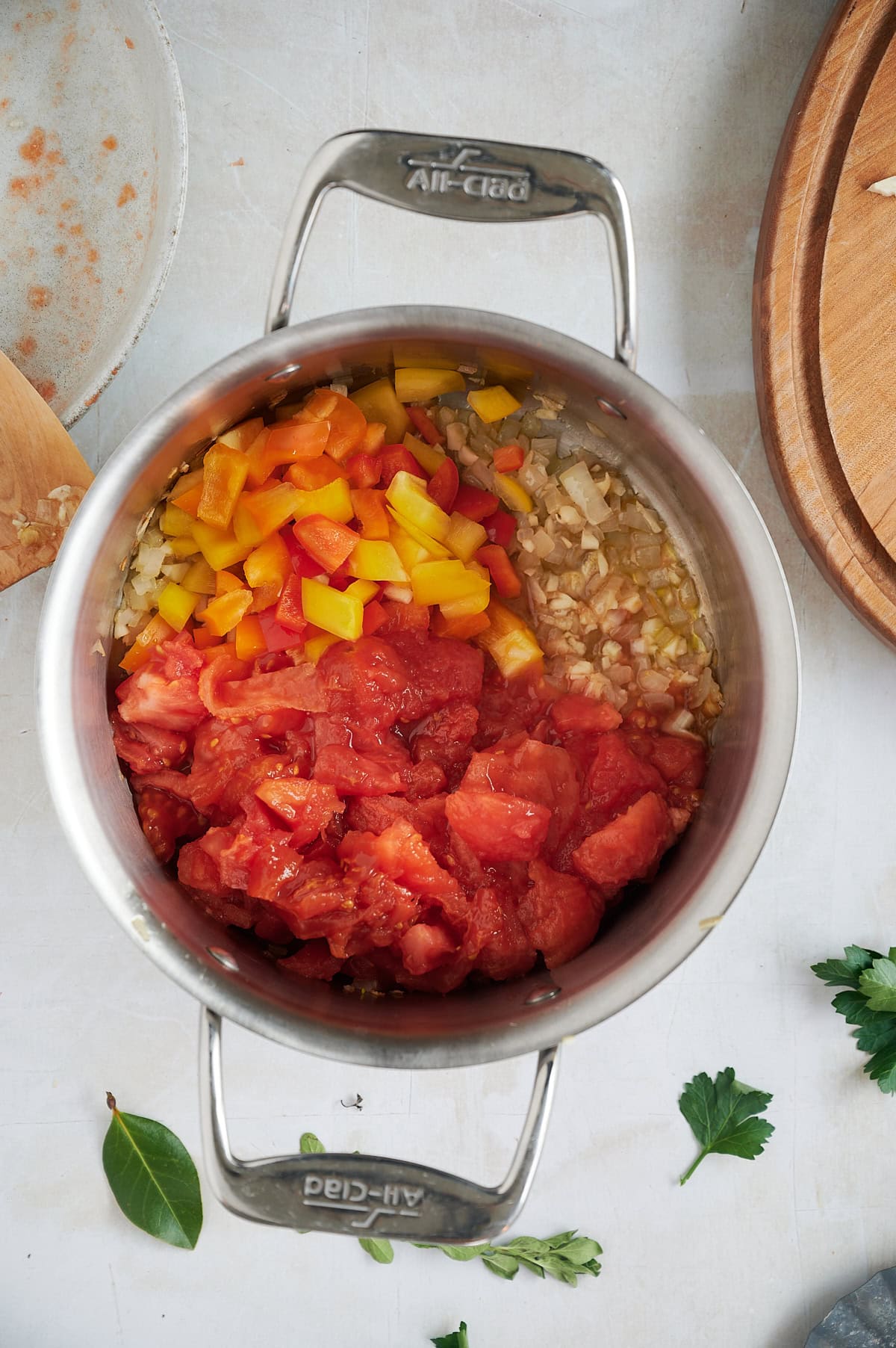 Three kinds of peppers with onions and garlic in a stainless soup pot with herbs on a cream background.