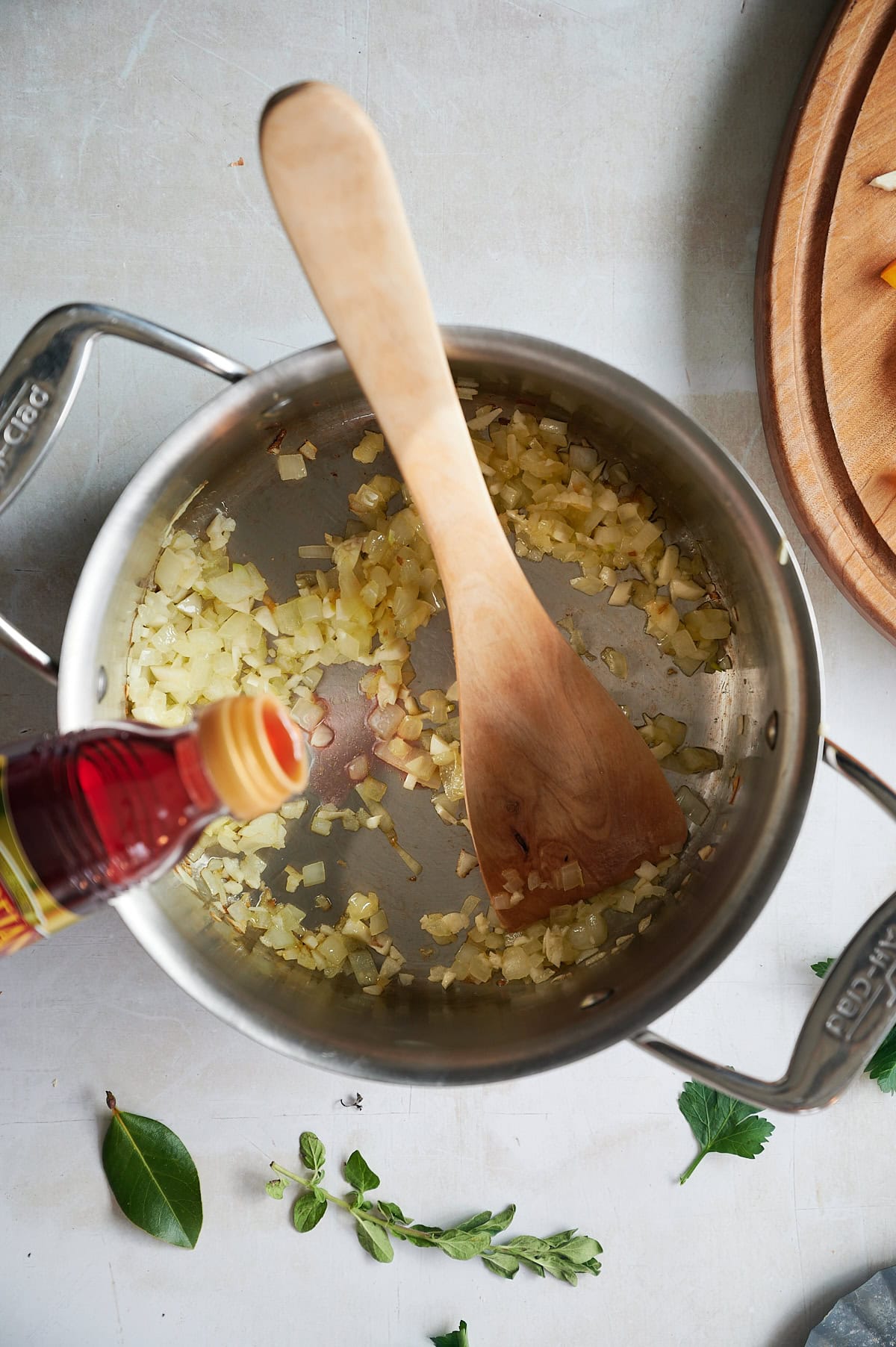 Red wine vinegar being poured into a stainless pot of sauteed onions and garlic being stirred by a wooden paddle.