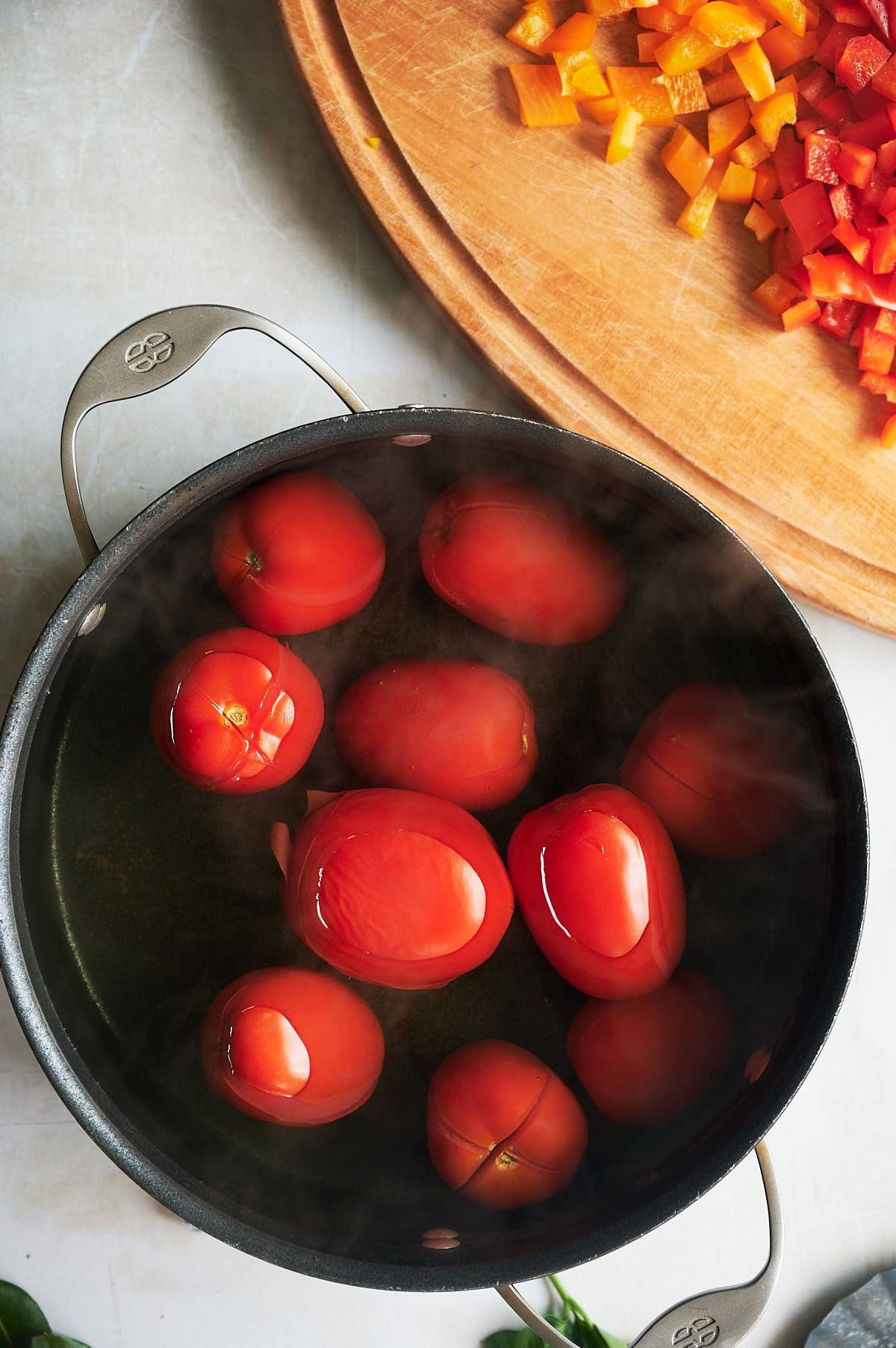 Pot of water covered tomatoes near a cutting board of peppers for stewed tomatoes.