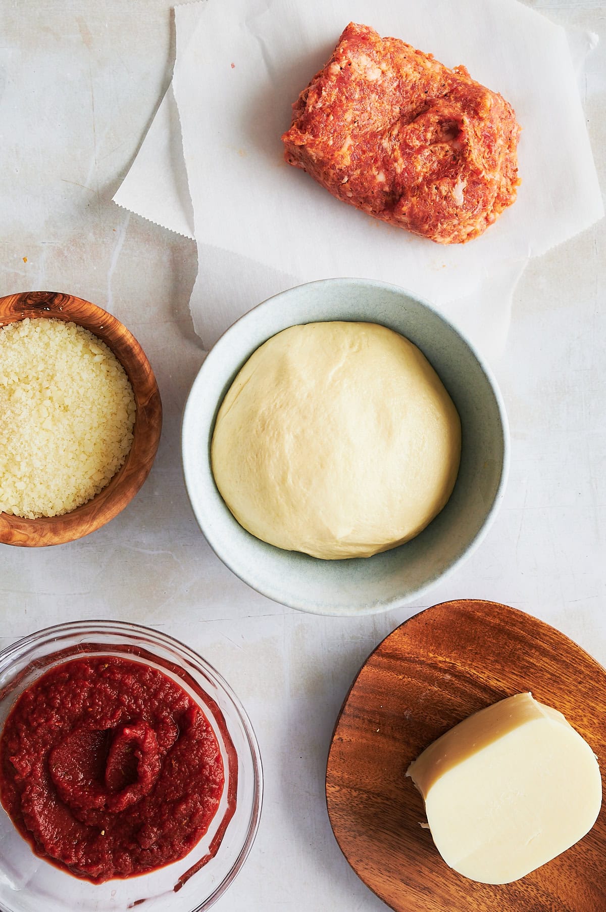 Ingredients for sausage pizza in bowls and on parchment including sauce, cheese and pizza dough.