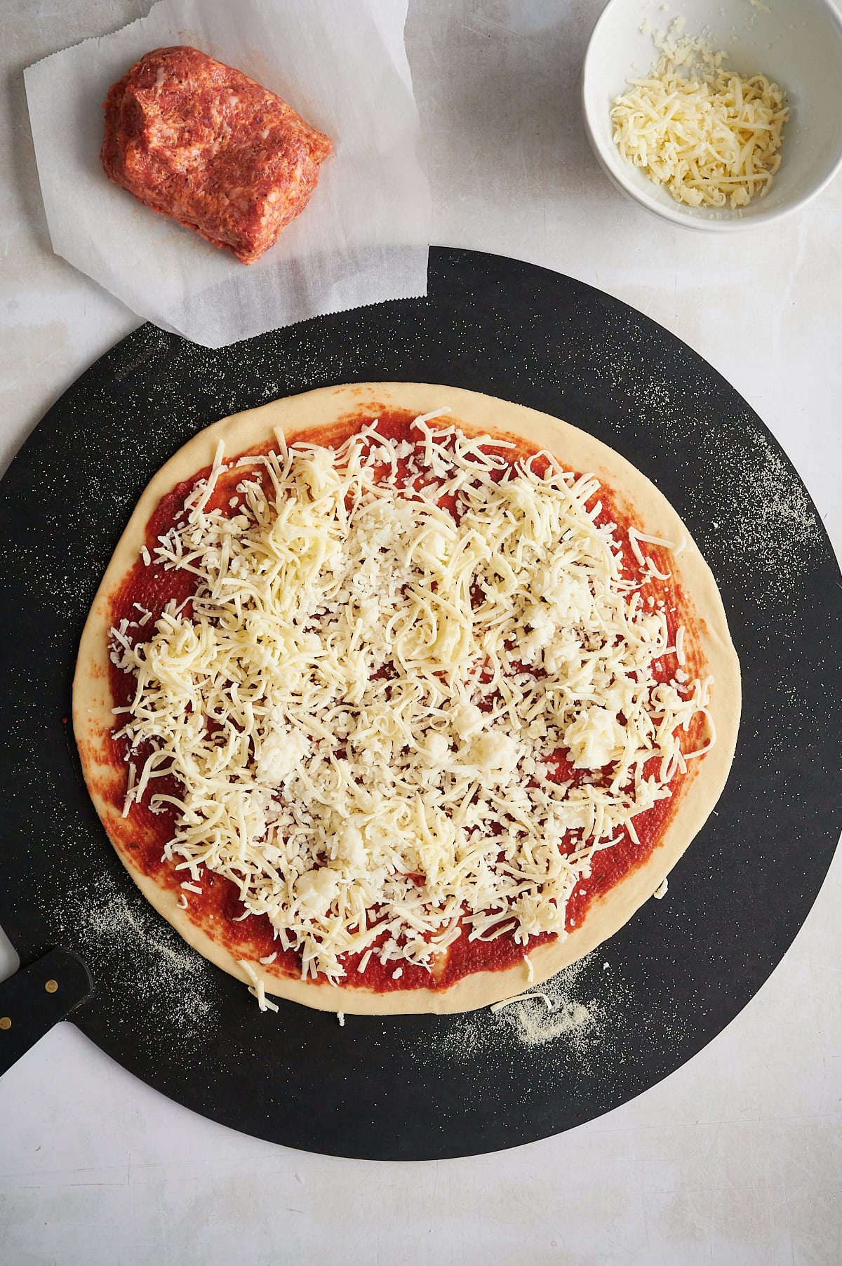 Unbaked cheese thin crust pizza on a brown pizza peel and wooden board with sausage and cheese nearby