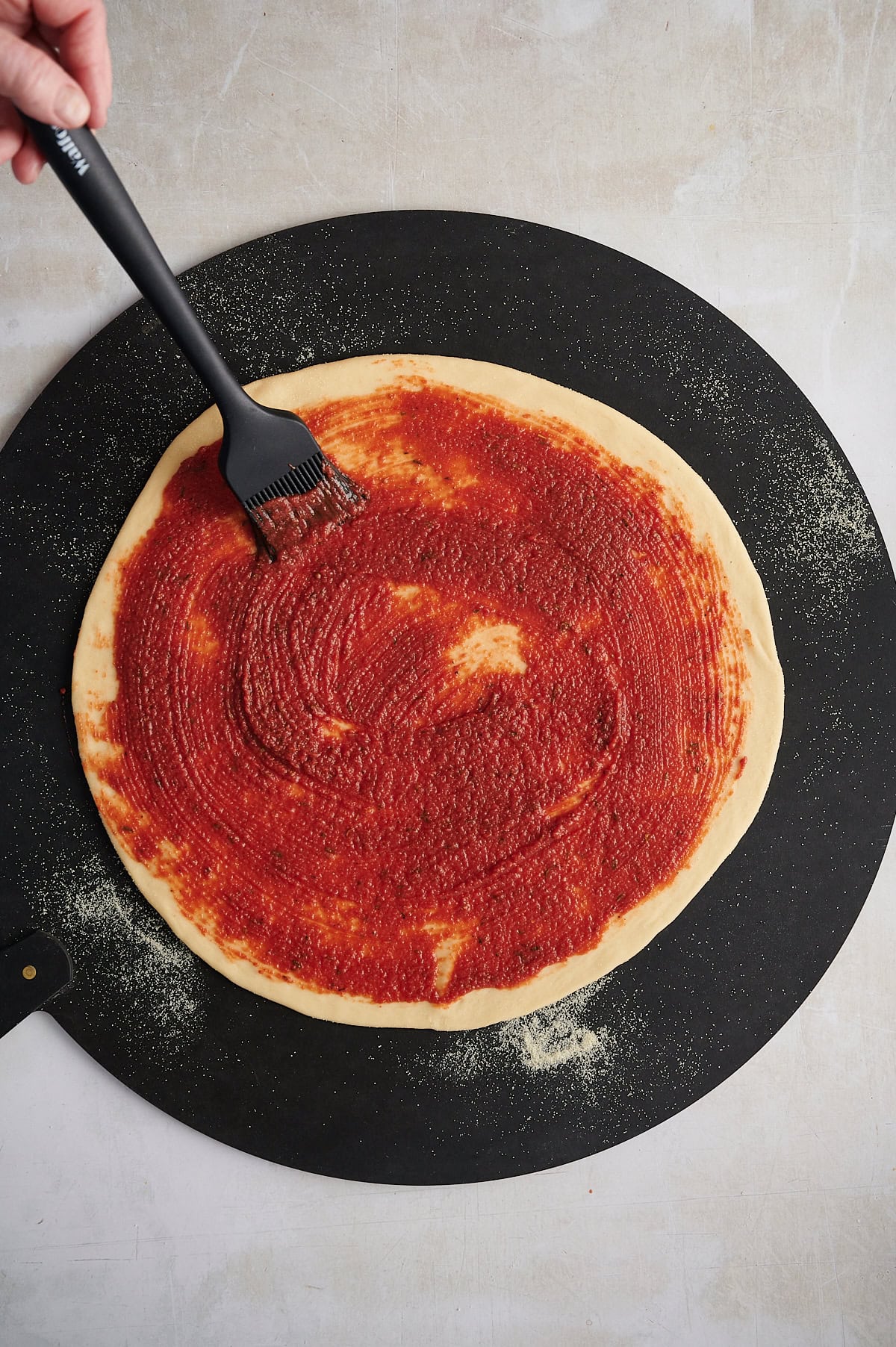 Hand brushing tomato sauce on pizza crust on black pizza stone.