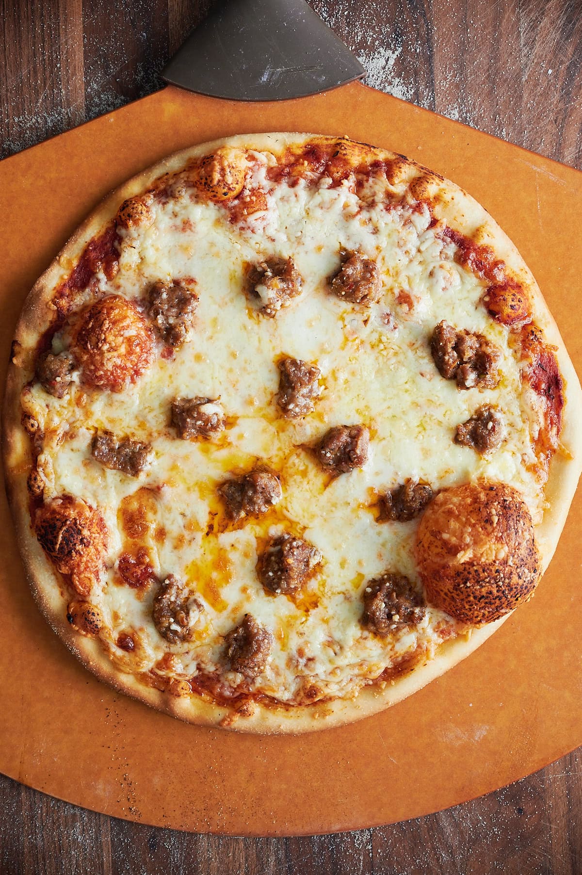Whole sausage and cheese thin crust pizza on a brown pizza peel and wooden board.