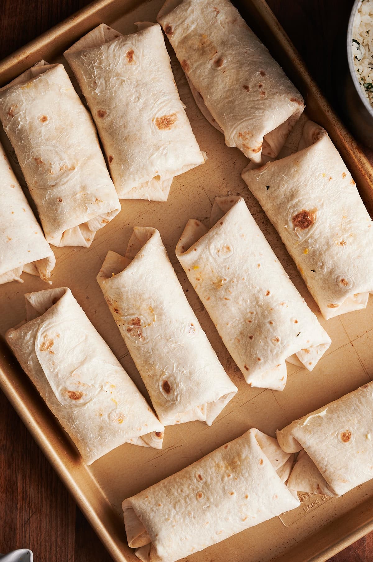 A baking tray filled with neatly arranged, baked beef burritos on a gold baking tray and wooden background.