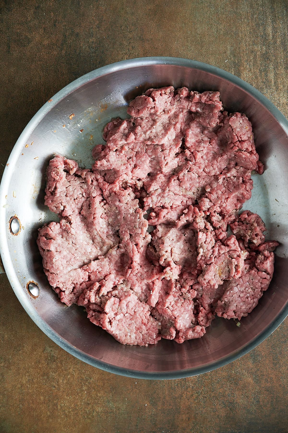 A stainless steel pan contains raw ground beef formed into rough, uneven sections sitting on a textured brown surface.