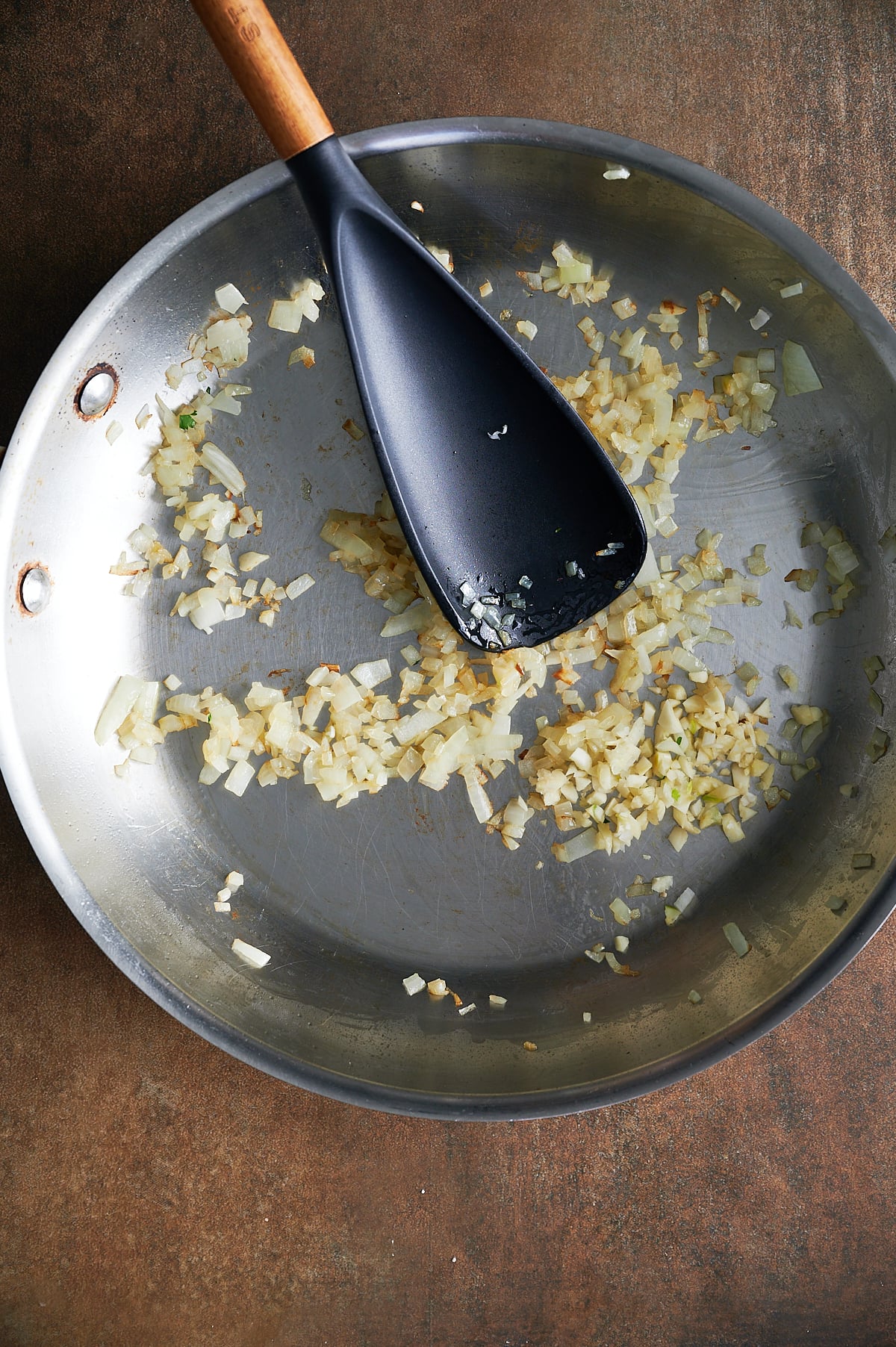Chopped golden browned onions and garlic sautéed in a stainless steel pan with a black spatula resting on top.