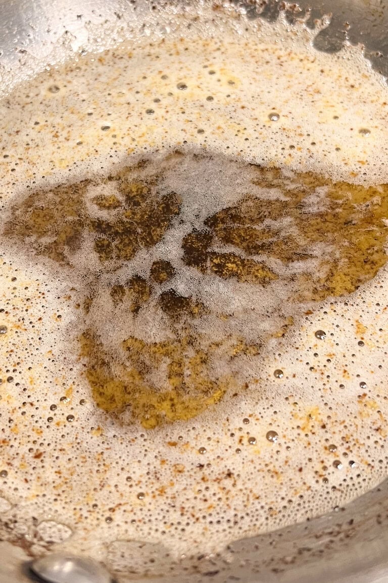 Close-up of bubbling browned butter in a pan, showing a mixture of light and dark brown specks with foam on top.