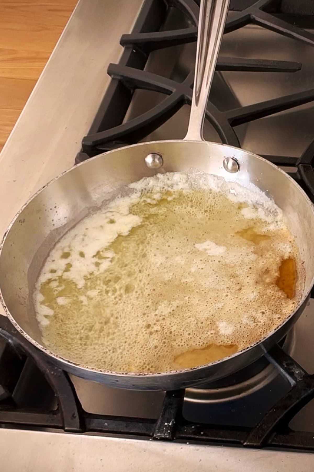 A stainless steel pan on a stove contains melted butter, turning golden brown with frothy bubbles.
