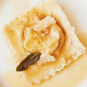 A single ravioli on a white plate, topped with shaved Parmesan cheese and crispy sage, drizzled with brown butter sauce.