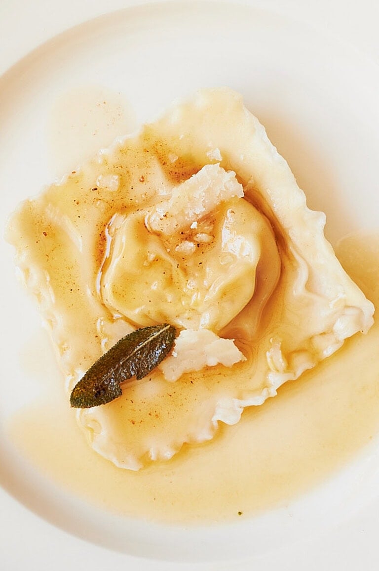 A single ravioli on a white plate, topped with shaved Parmesan cheese and crispy sage, drizzled with brown butter sauce.