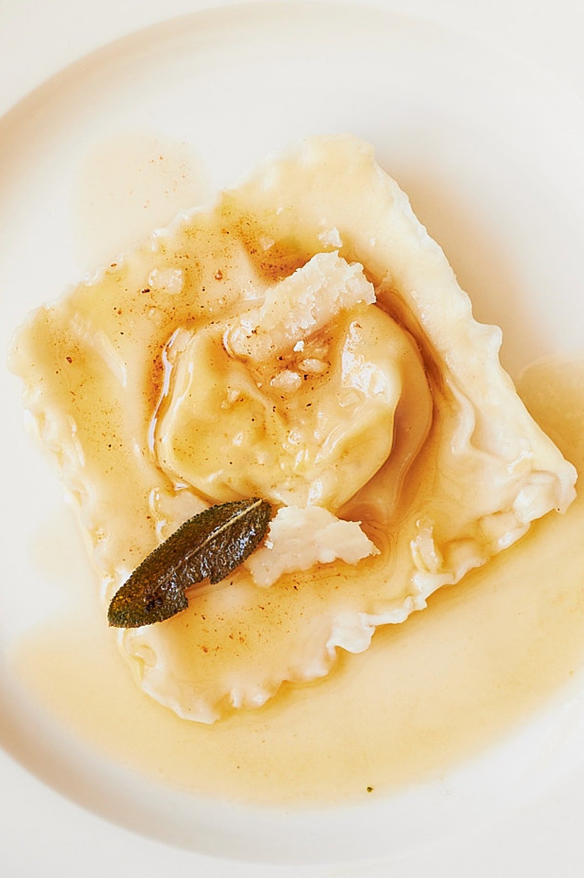 A single ravioli on a white plate, topped with shaved Parmesan cheese and crispy sage, drizzled with brown butter sauce.