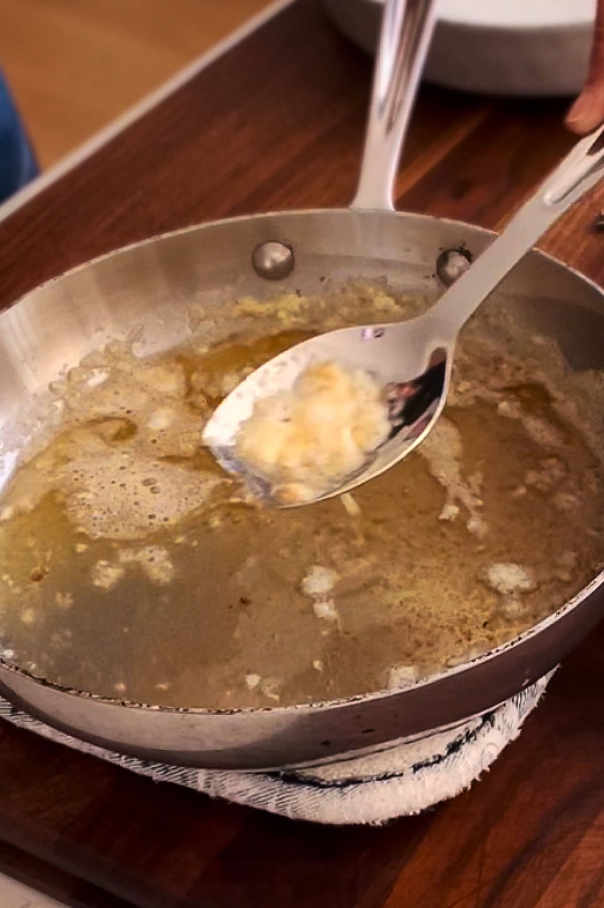 A stainless steel spoon is scooping out a milk solids from melted brown butter food or residue. A cloth is placed underneath the pan for stability.
