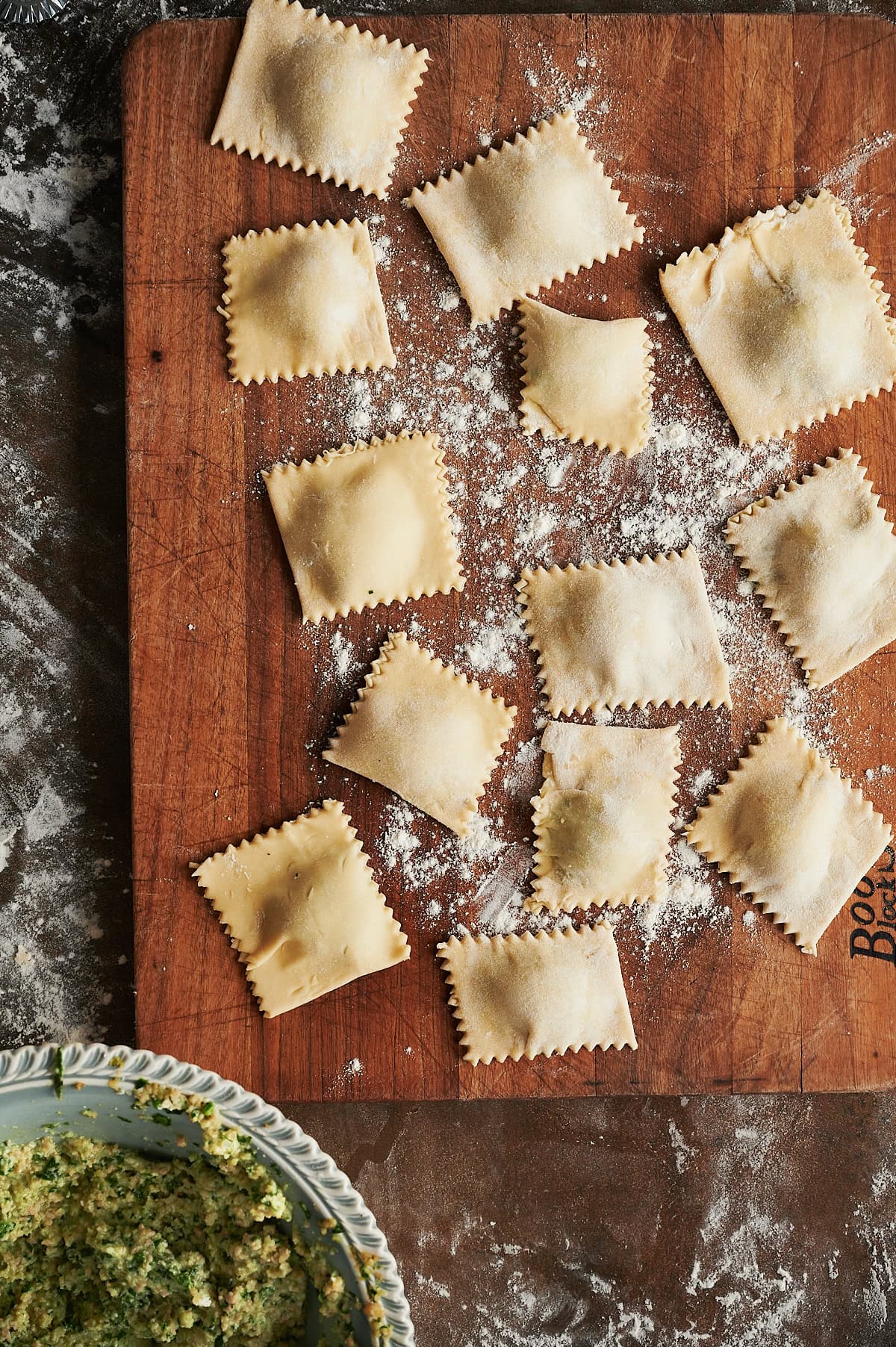 Wooden cutting board with cut uncooked ravioli sprinkled with flour and gray bowl of filling nearby.