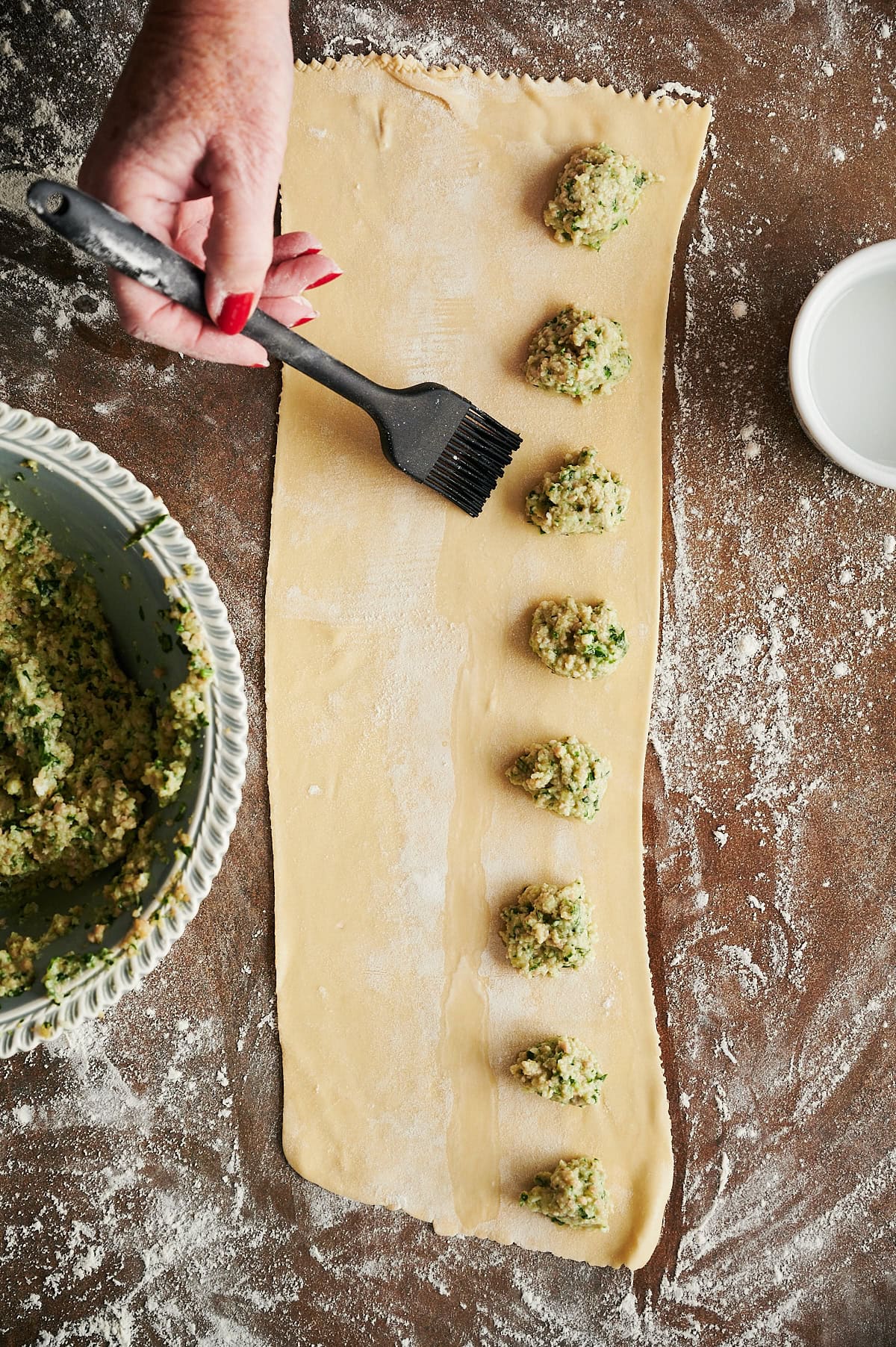 Person brushing water around chicken ravioli filling on ravioli dough with a brown background and bowl of filling nearby.