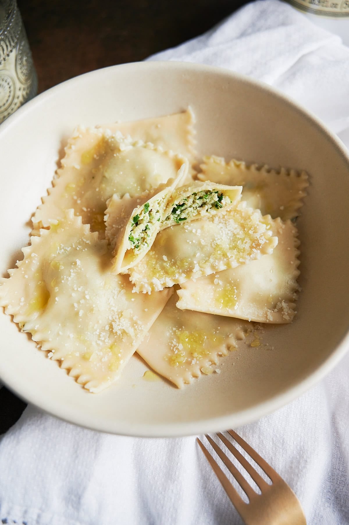 Table setting of a white bowl of chicken ravioli drizzled with olive oil and parmesan cheese with a fork on a white napkin nearby.