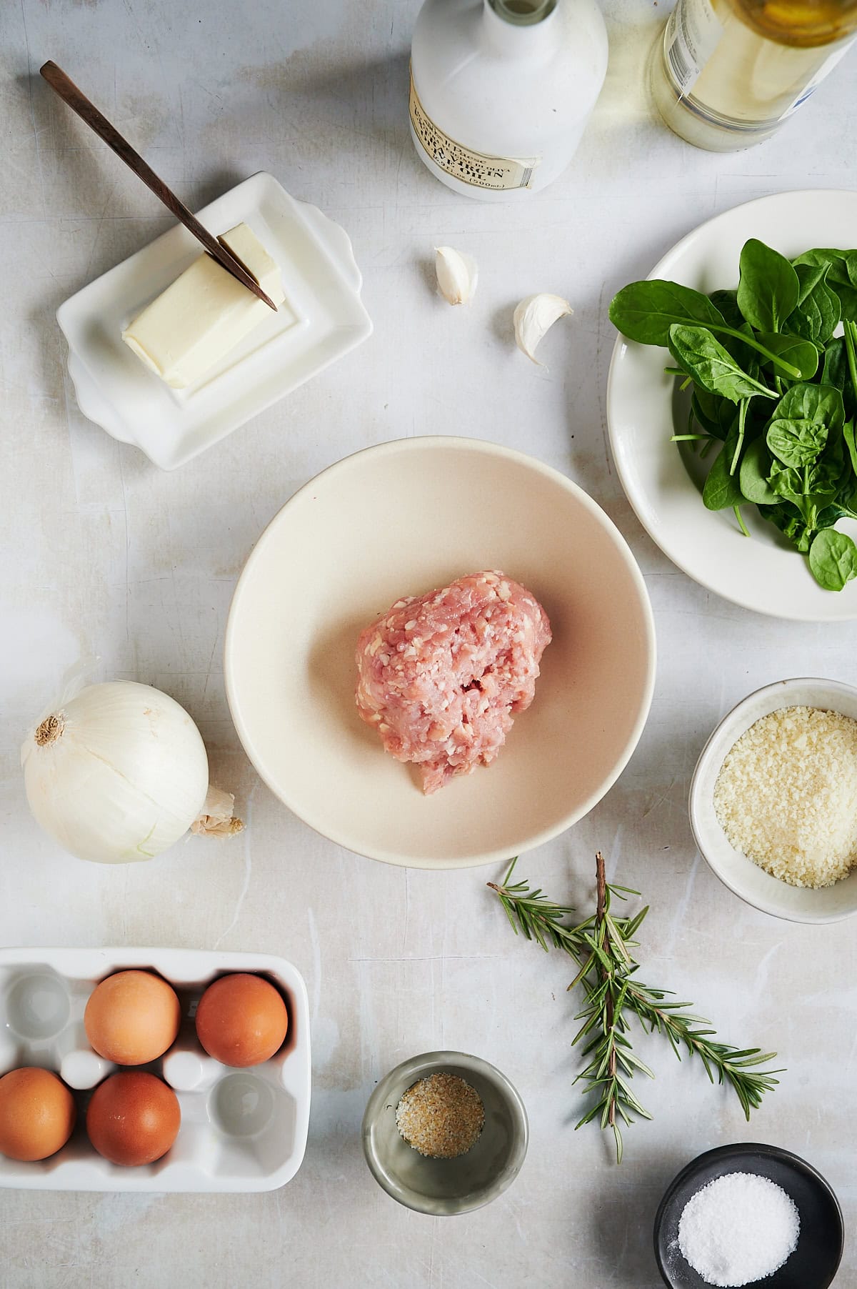 Ingredients on a table including minced meat in a bowl, spinach, eggs, a white onion, butter, minced garlic, grated cheese, and herbs with bottles of oil and a small bowl of salt and pepper are also visible.