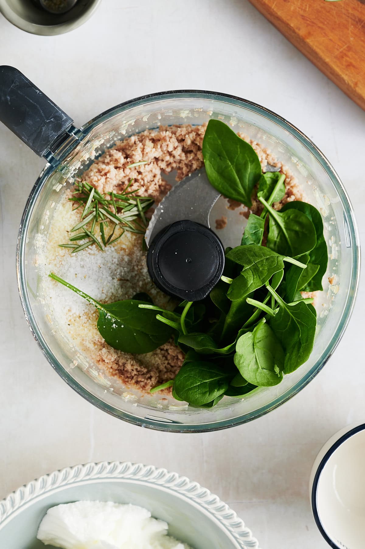 A food processor bowl containing minced chicken, fresh spinach leaves, rosemary sprigs, and a sprinkle of salt for ravioli.