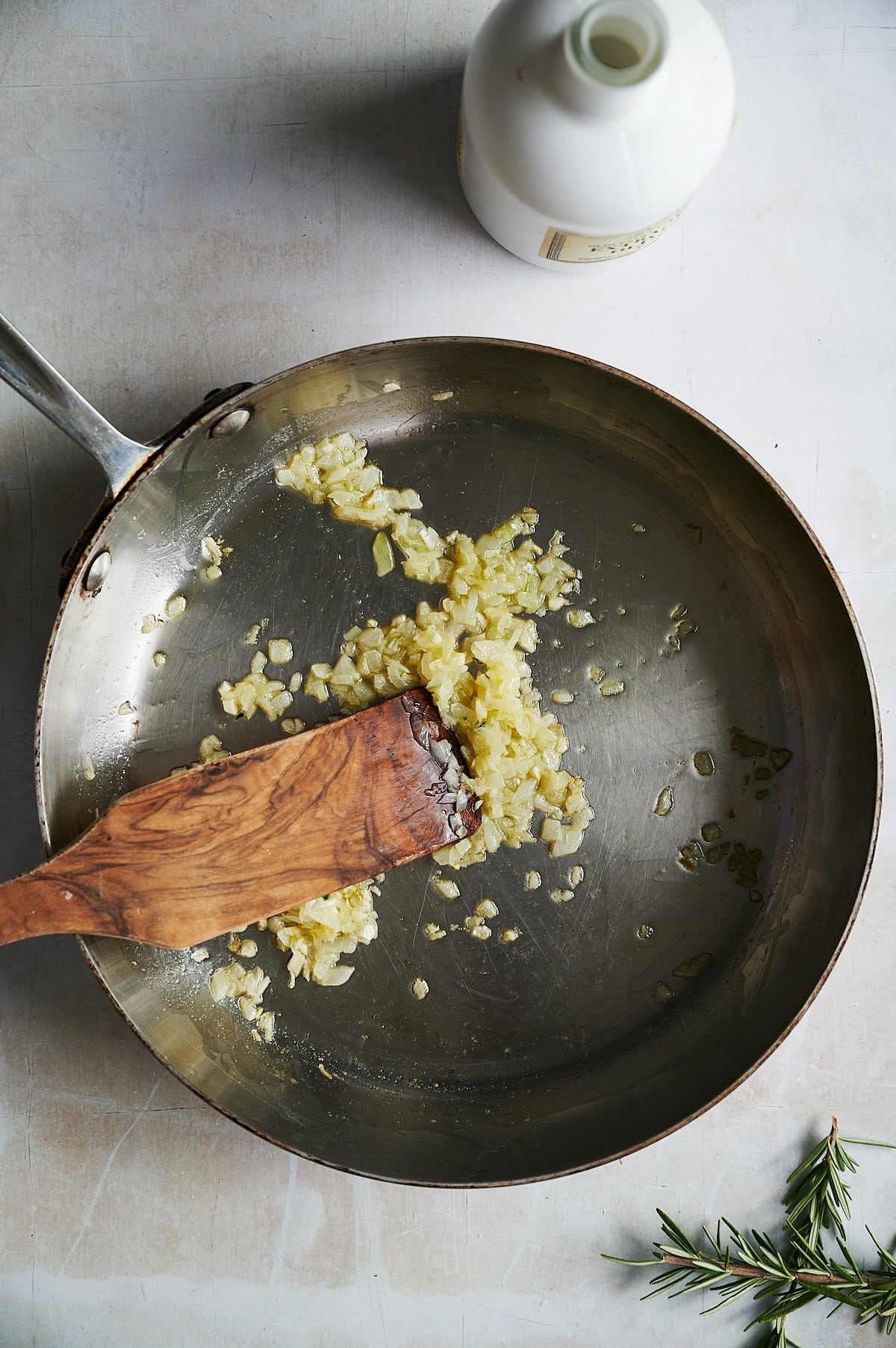 A frying pan with sautéed shallots being stirred by a wooden spatula on a light-colored surface with a bottle and a sprig of rosemary placed nearby.