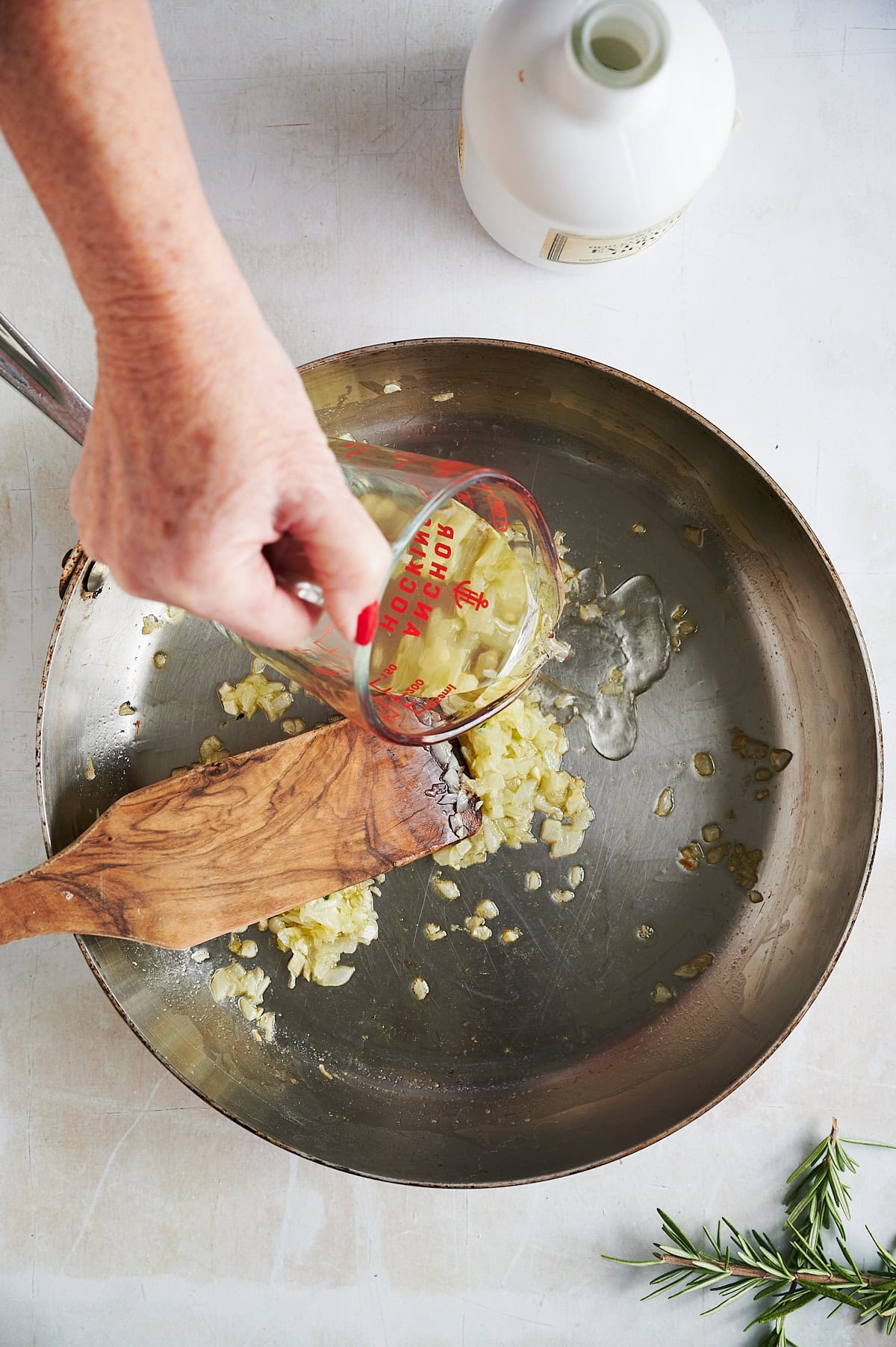 A person pouring liquid from a measuring cup into a skillet with chopped onions and a wooden spatula is in the skillet, and a small sprig of rosemary is nearby.