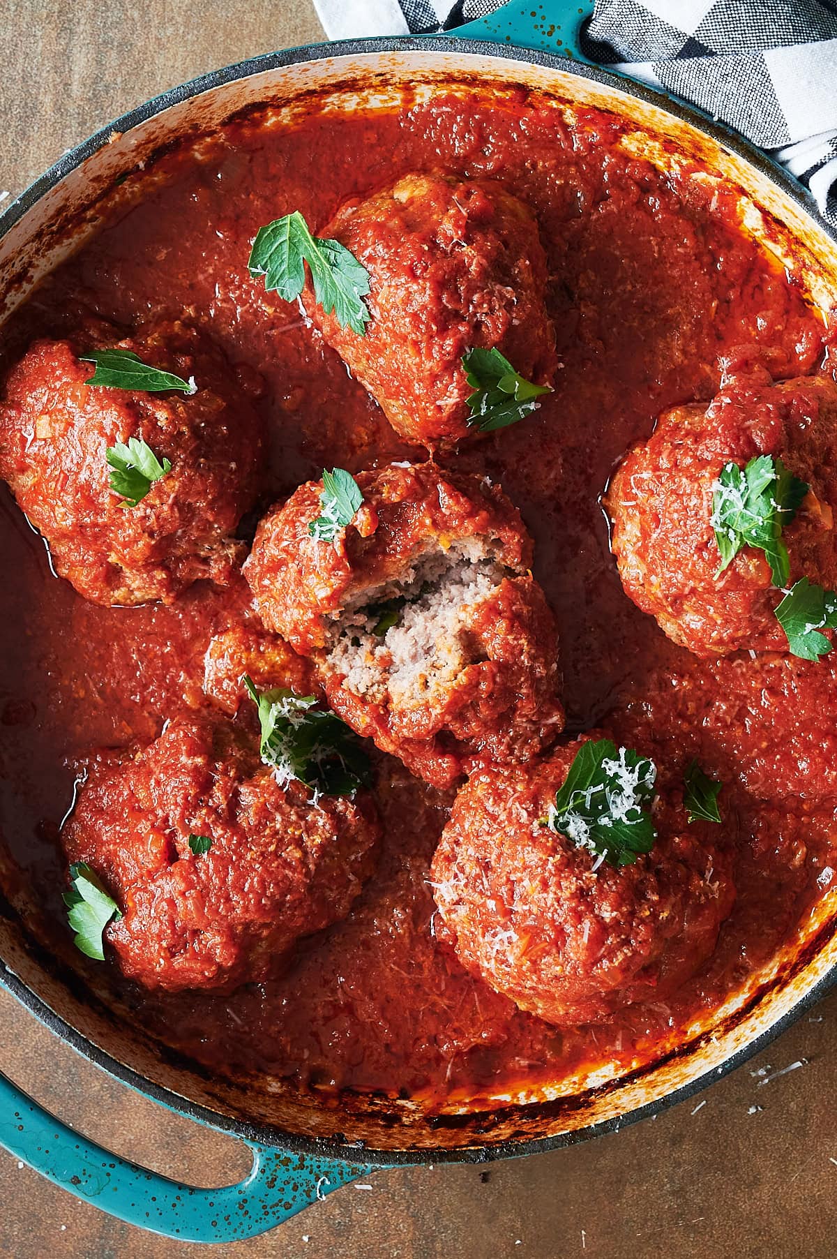 A close-up of a blue pot filled with large meatballs covered in tomato sauce and garnished with fresh parsley and grated cheese. One meatball is split open, revealing the inside.