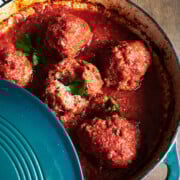 A blue enameled pot filled with large meatballs in rich tomato sauce with one meatball cut open. Fresh parsley garnishes the dish and a checkered cloth is nearby.