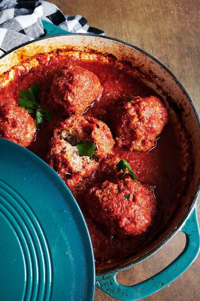 A blue enameled pot filled with large meatballs in rich tomato sauce with one meatball cut open. Fresh parsley garnishes the dish and a checkered cloth is nearby.
