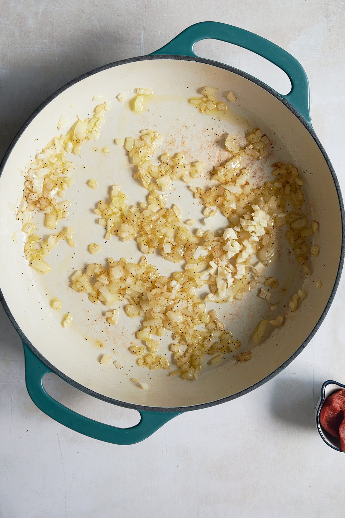 Chopped onions and garlic sautéing in a large white and teal enameled pot on a light, textured surface.
