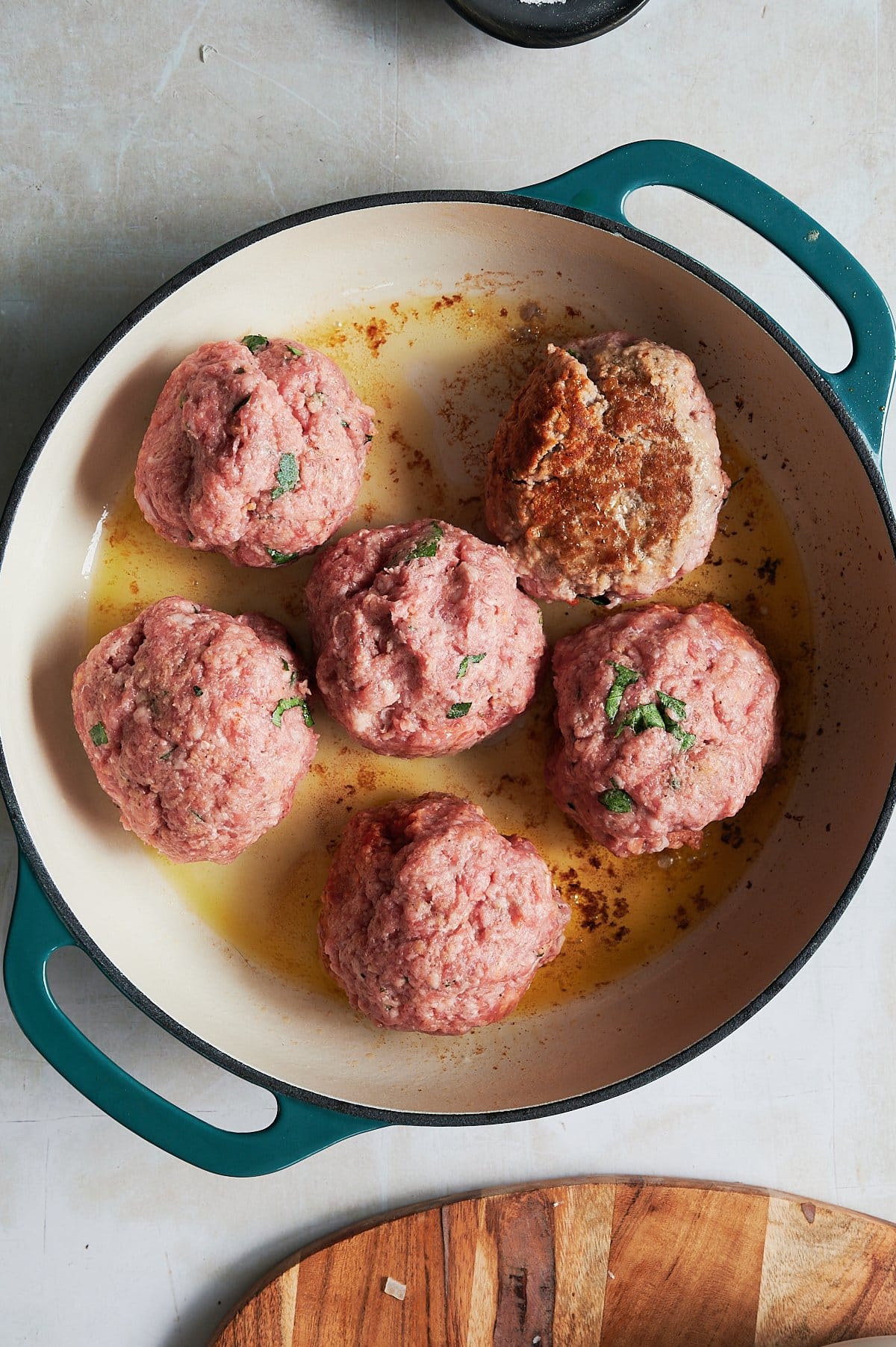 A round dish containing five giant, raw meatballs, one partially browned, with herbs sprinkled on top.