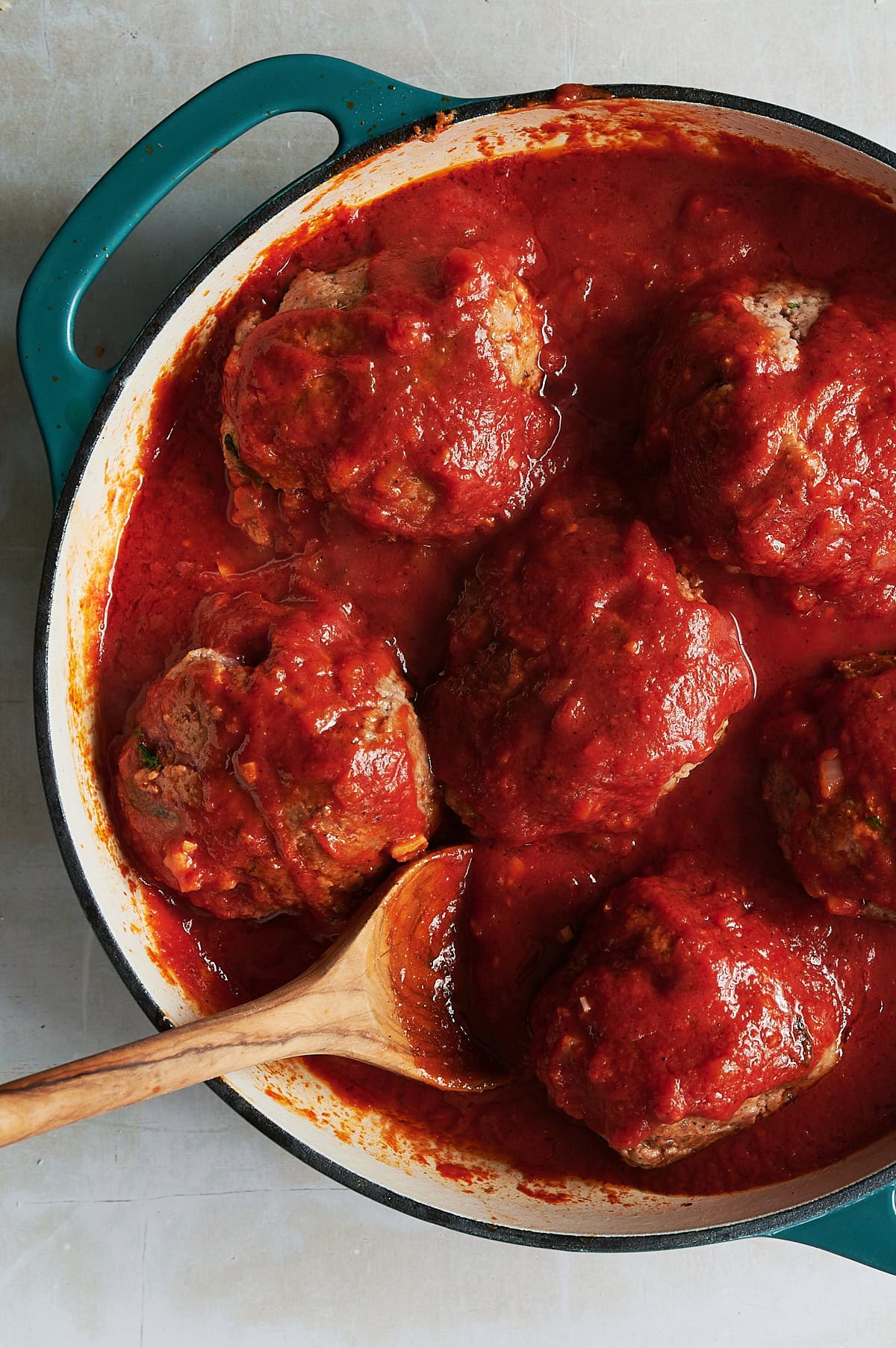 A teal-handled pot filled with large meatballs covered in rich red tomato sauce with a wooden spoon resting on the side of the pot.