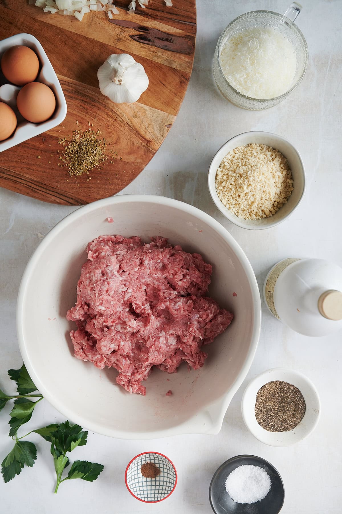 Ingredients for meatballs are laid out on a surface, including a bowl of ground meat, eggs, breadcrumbs, grated cheese, garlic, parsley, onion, salt, pepper, and spices.