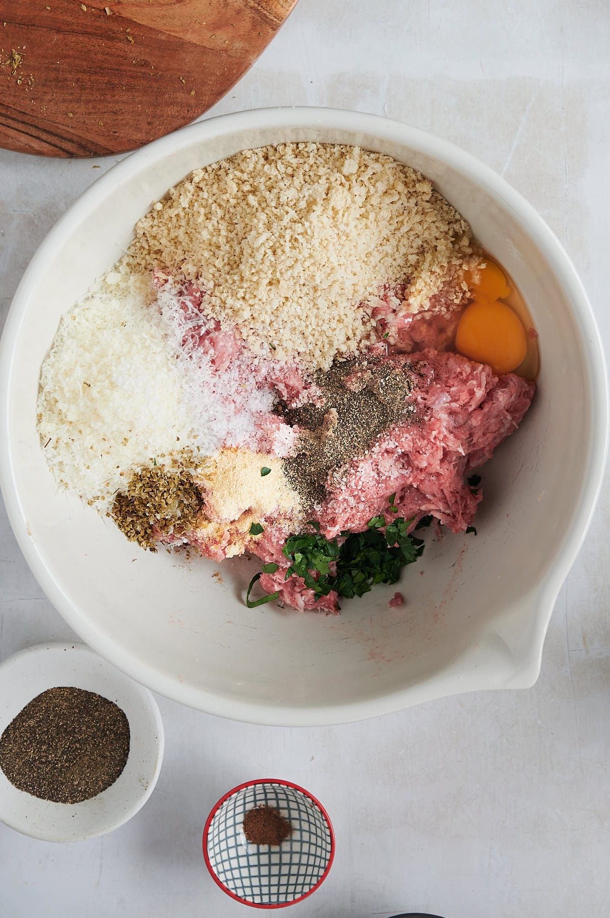 A large white bowl containing ingredients for giant meatballs, including ground meat, breadcrumbs, grated cheese, an egg, chopped parsley, and various spices. Nearby are small bowls with additional spices and a wooden chopping board nearby.