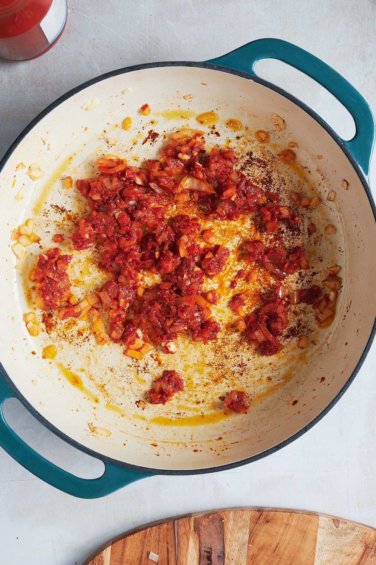 A white pan with teal handles contains sautéed onions and tomatoes, creating a rich red mixture with oily textures. The pan sits on a light surface, and a glimpse of a wooden board and a red container is visible nearby.