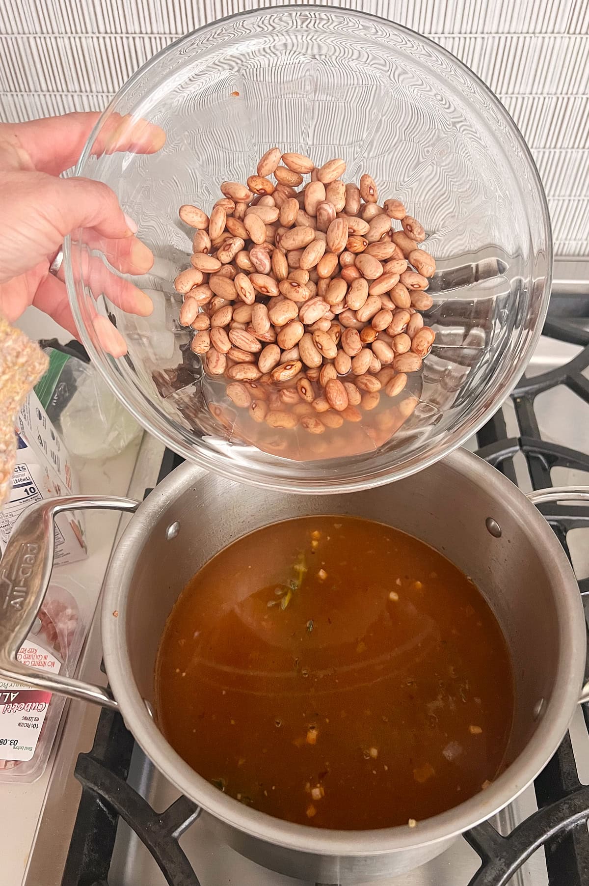 A hand holds a glass bowl filled with cranberry beans above a pot of simmering brown broth on a stovetop.