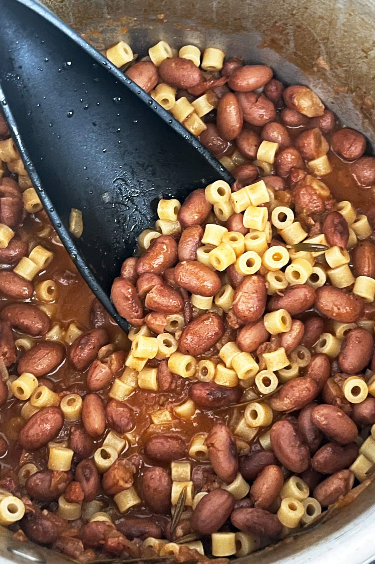 A pot of pasta e fagioli with small pasta tubes and kidney beans in a tomato-based sauce and a black spoon stirring the mixture.