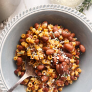 A gray bowl of pasta e fagioli, brimming with cranberry beans and topped with grated cheese and fresh herbs and a fork resting invitingly in the bowl.