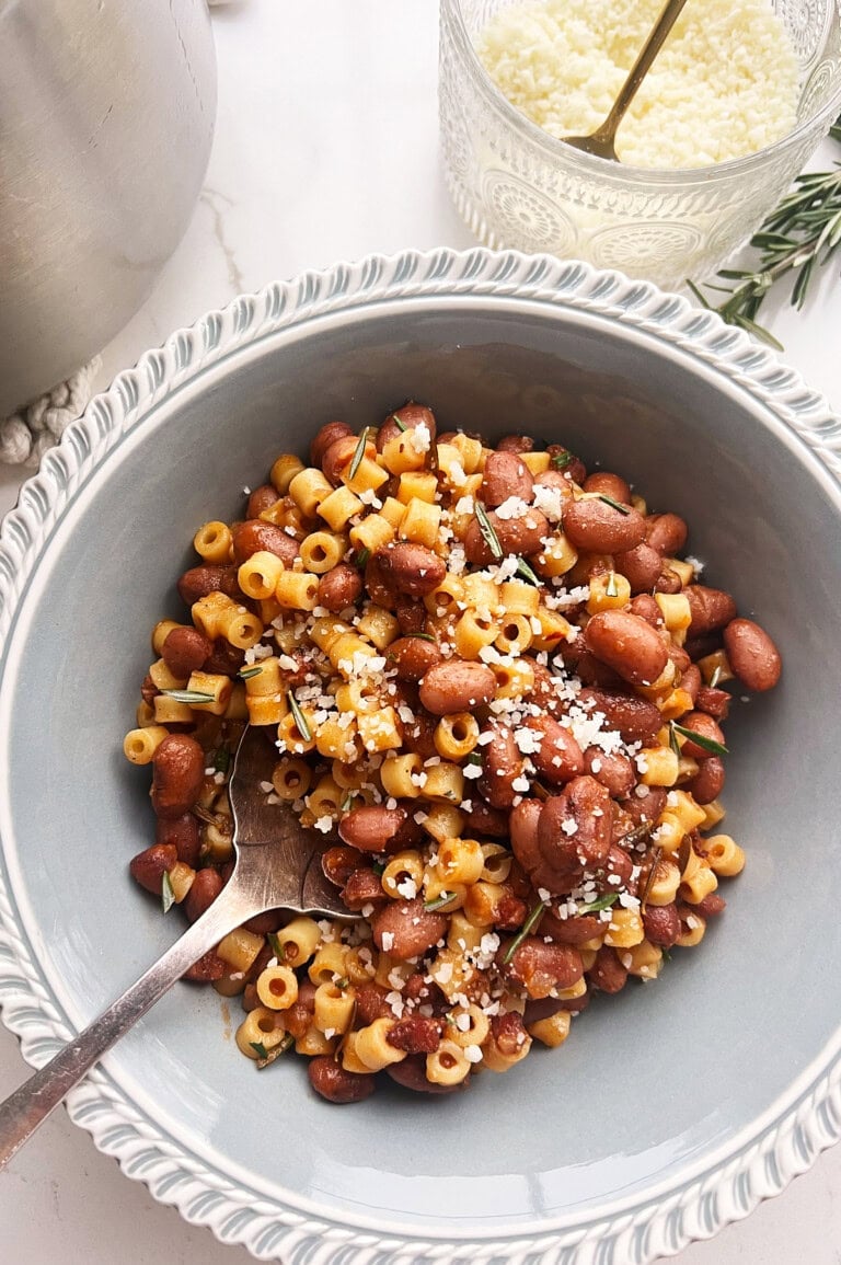 A gray bowl of pasta e fagioli, brimming with cranberry beans and topped with grated cheese and fresh herbs and a fork resting invitingly in the bowl.