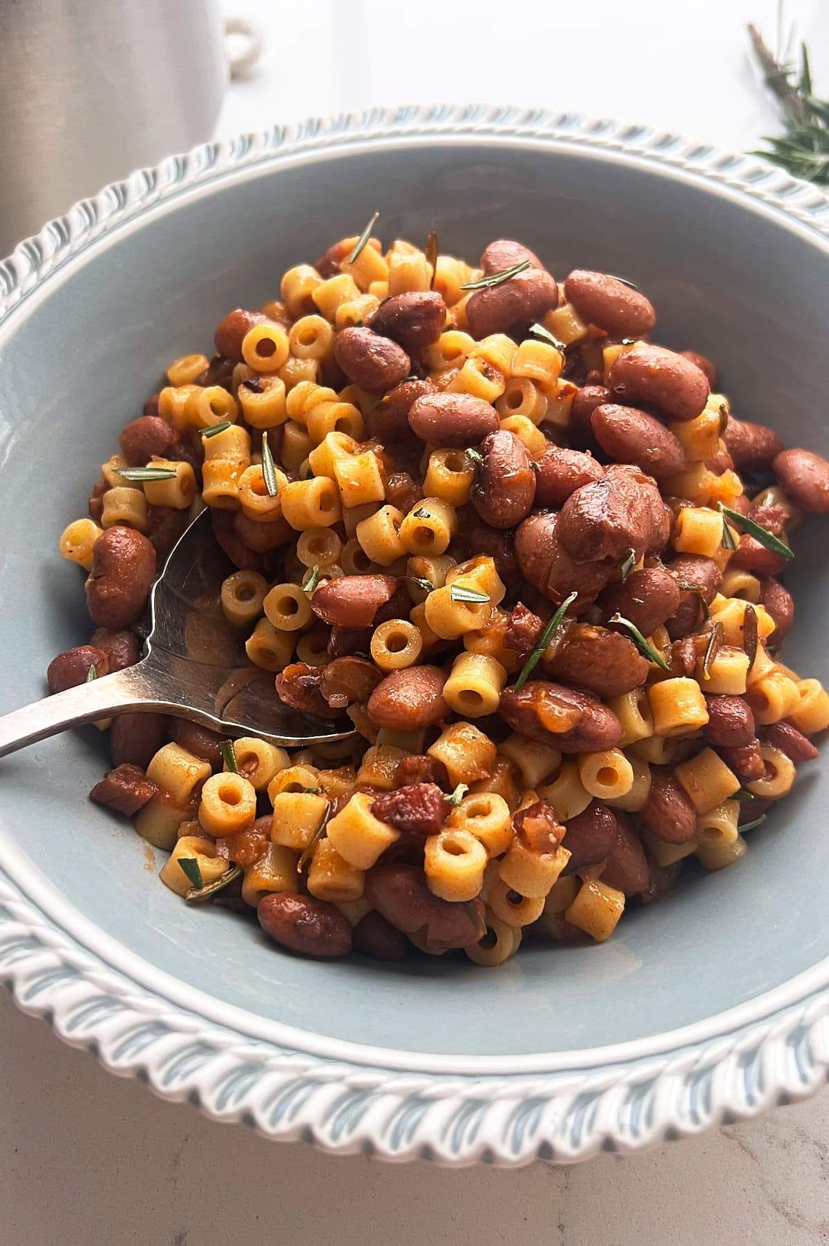 A bowl of pasta topped with kidney beans and garnished with herbs. A spoon rests in the ceramic bowl, and the combination appears to be savory, featuring round pasta pieces and red beans. The edge of a pot is visible in the background.